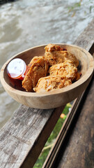 Tahu walik. Crispy browned tofu walik served in a natural wooden bowl with spicy chili sauce, with a blurry river flowing in the background. Menu at an outdoor cafe.