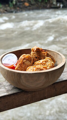Tahu walik. Crispy browned tofu walik served in a natural wooden bowl with spicy chili sauce, with a blurry river flowing in the background. Menu at an outdoor cafe.