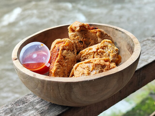 Tahu walik. Crispy browned tofu walik served in a natural wooden bowl with spicy chili sauce, with a blurry river flowing in the background. Menu at an outdoor cafe.