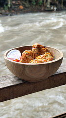 Tahu walik. Crispy browned tofu walik served in a natural wooden bowl with spicy chili sauce, with a blurry river flowing in the background. Menu at an outdoor cafe.