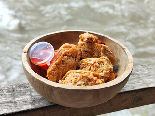 Tahu walik. Crispy browned tofu walik served in a natural wooden bowl with spicy chili sauce, with a blurry river flowing in the background. Menu at an outdoor cafe.