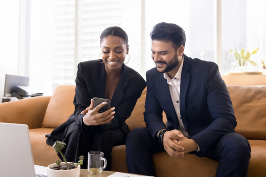 Casual collaboration and modern tech. Smiling young businesswoman sit on couch in office show photo video to male colleague on smartphone screen share creative idea funny business meme at social media