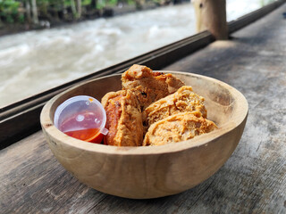 Tahu walik. Crispy browned tofu walik served in a natural wooden bowl with spicy chili sauce, with a blurry river flowing in the background. Menu at an outdoor cafe.