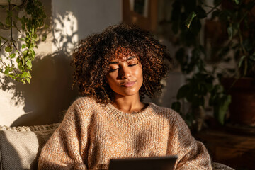 Curly Haired Black Woman Wearing Cozy Beige Sweater Enjoying Tablet Amid Sunlit Indoor Greenery Serenity Concept. Relaxed Woman