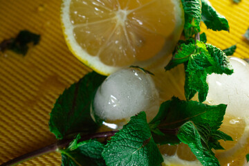 Refreshing summer close-up of lemon slices, mint leaves, and round ice cubes on a yellow...
