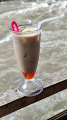 A tall glass of cold chocolate milk with ice cubes and a straw, placed on the edge of a weathered wooden fence with a blurry backdrop of a fast flowing river. Outdoor cafe.