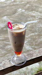 A tall glass of cold chocolate milk with ice cubes and a straw, placed on the edge of a weathered wooden fence with a blurry backdrop of a fast flowing river. Outdoor cafe.