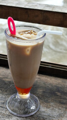 A tall glass of cold chocolate milk with ice cubes and a straw, placed on the edge of a weathered wooden fence with a blurry backdrop of a fast flowing river. Outdoor cafe.