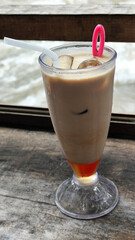 A tall glass of cold chocolate milk with ice cubes and a straw, placed on the edge of a weathered wooden fence with a blurry backdrop of a fast flowing river. Outdoor cafe.