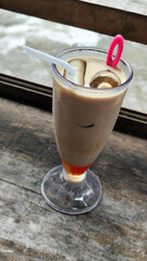 A tall glass of cold chocolate milk with ice cubes and a straw, placed on the edge of a weathered wooden fence with a blurry backdrop of a fast flowing river. Outdoor cafe.