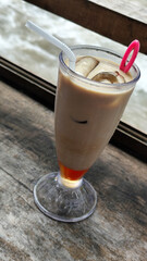 A tall glass of cold chocolate milk with ice cubes and a straw, placed on the edge of a weathered wooden fence with a blurry backdrop of a fast flowing river. Outdoor cafe.