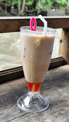 A tall glass of cold chocolate milk with ice cubes and a straw, placed on the edge of a weathered wooden fence with a blurry backdrop of a fast flowing river. Outdoor cafe.