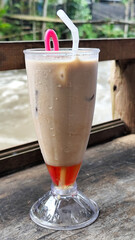 A tall glass of cold chocolate milk with ice cubes and a straw, placed on the edge of a weathered wooden fence with a blurry backdrop of a fast flowing river. Outdoor cafe.