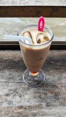 A tall glass of cold chocolate milk with ice cubes and a straw, placed on the edge of a weathered wooden fence with a blurry backdrop of a fast flowing river. Outdoor cafe.