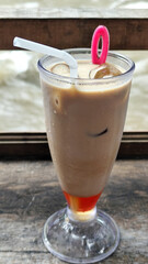A tall glass of cold chocolate milk with ice cubes and a straw, placed on the edge of a weathered wooden fence with a blurry backdrop of a fast flowing river. Outdoor cafe.