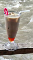 A tall glass of cold chocolate milk with ice cubes and a straw, placed on the edge of a weathered wooden fence with a blurry backdrop of a fast flowing river. Outdoor cafe.