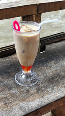 A tall glass of cold chocolate milk with ice cubes and a straw, placed on the edge of a weathered wooden fence with a blurry backdrop of a fast flowing river. Outdoor cafe.