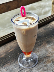A tall glass of cold chocolate milk with ice cubes and a straw, placed on the edge of a weathered wooden fence with a blurry backdrop of a fast flowing river. Outdoor cafe.