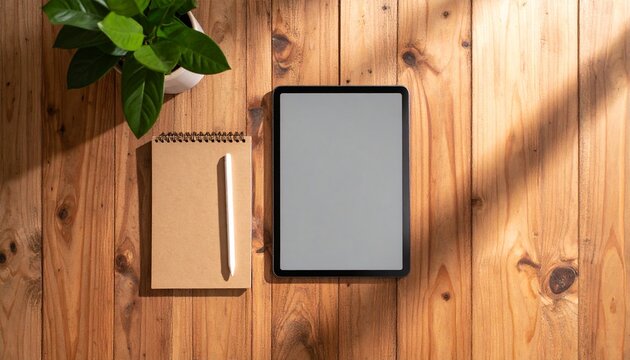 Wooden table surface with digital tablet, notepad, pen, and plant.