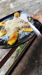 A plate of grilled bananas sprinkled with grated cheese and sweetened condensed milk on a black plate, with a backdrop of a rocky river and blurry green trees. Outdoor cafe.