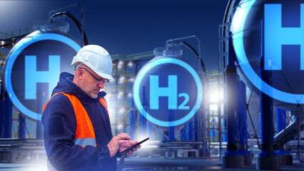 Engineer in helmet and vest uses tablet at hydrogen plant at night with large H2 tanks in...