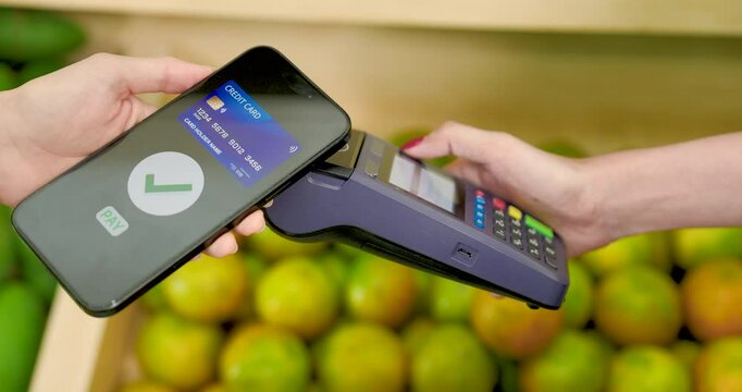 Asian female adult making cashless payment by tapping smartphone on terminal for purchase inside tropical fruit store during digital checkout in small business retail setting