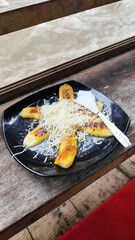A plate of grilled bananas sprinkled with grated cheese and sweetened condensed milk on a black plate, with a backdrop of a rocky river and blurry green trees. Outdoor cafe.
