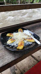 A plate of grilled bananas sprinkled with grated cheese and sweetened condensed milk on a black plate, with a backdrop of a rocky river and blurry green trees. Outdoor cafe.