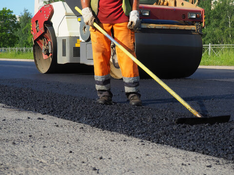 Worker leveling asphalt with hand tool. Road construction. Laying asphalt on city streets. Roller asphalt paver under summer sky. Cropped contractor in uniform building road. Highway construction