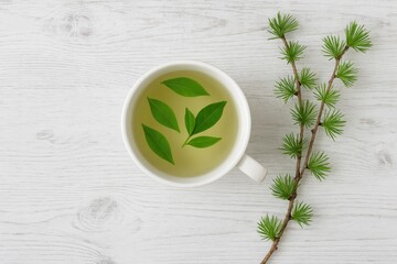 A white cup filled with floating green tea leaves, set against a rustic wooden backdrop with a larch branch for added charm.
