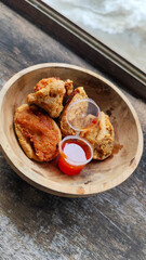 Tahu walik. Crispy browned tofu walik served in a natural wooden bowl with spicy chili sauce, with a blurry river flowing in the background. Menu at an outdoor cafe.
