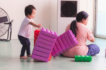 Mother and son enjoying playtime with building blocks. A two-year-old boy is secretly pushing a plastic block down onto the ground behind his mother.