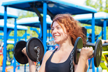 Smiling red-haired curly slim woman in sportswear exercising with dumbbells.