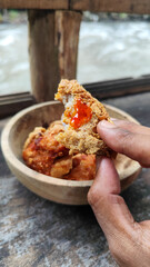 Tahu walik. Crispy browned tofu walik served in a natural wooden bowl with spicy chili sauce, with a blurry river flowing in the background. Menu at an outdoor cafe.
