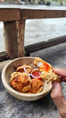 Tahu walik. Crispy browned tofu walik served in a natural wooden bowl with spicy chili sauce, with a blurry river flowing in the background. Menu at an outdoor cafe.