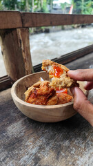 Tahu walik. Crispy browned tofu walik served in a natural wooden bowl with spicy chili sauce, with a blurry river flowing in the background. Menu at an outdoor cafe.