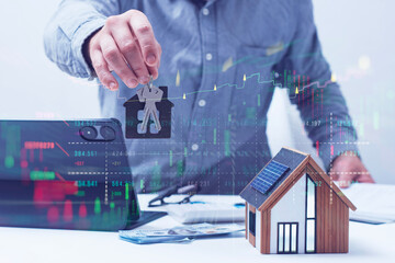 Businessman holds house shaped keychain with keys over a desk with a house model, tablet, and dollar bills with a digital overlay of stock market charts and an upward trend line.