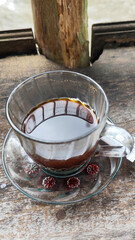 A cup of hot black coffee in a glass cup with matching saucer and spoon, placed on the edge of a weathered wooden fence with a backdrop of a flowing river and blurry green trees. Outdoor cafe menu.