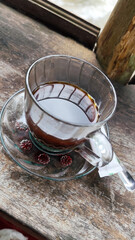 A cup of hot black coffee in a glass cup with matching saucer and spoon, placed on the edge of a weathered wooden fence with a backdrop of a flowing river and blurry green trees. Outdoor cafe menu.