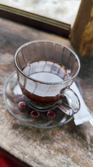 A cup of hot black coffee in a glass cup with matching saucer and spoon, placed on the edge of a weathered wooden fence with a backdrop of a flowing river and blurry green trees. Outdoor cafe menu.
