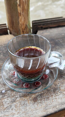 A cup of hot black coffee in a glass cup with matching saucer and spoon, placed on the edge of a weathered wooden fence with a backdrop of a flowing river and blurry green trees. Outdoor cafe menu.