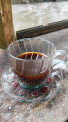A cup of hot black coffee in a glass cup with matching saucer and spoon, placed on the edge of a weathered wooden fence with a backdrop of a flowing river and blurry green trees. Outdoor cafe menu.