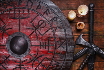 Old long medieval two handed knight warrior sword, battle axe and red wooden shield with runes on ancient table background.