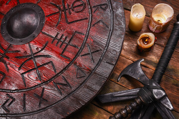 Old long medieval two handed knight warrior sword, battle axe and red wooden shield with runes on ancient table background.