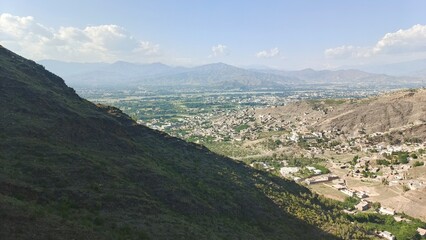 Serene Mountain Valley Landscape: Panoramic View of Village Nestled Amongst Lush Greenery and Majestic Mountains Under a Partly Cloudy Sky.