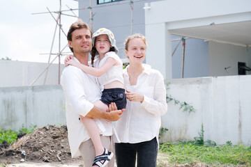 Father and mother are hugging their smiling daughter as they stand in front of the house.