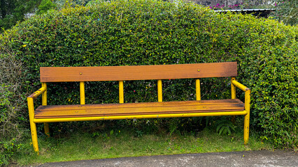 This image features a rustic park bench with a yellow metal frame and wooden slats, placed in front...