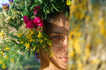 A girl of Slavic appearance with a wreath of wild flowers on her head looks out from behind a tree