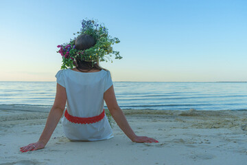 A girl of Slavic appearance with a wreath of wild flowers on her head sits on the shore of a lake....