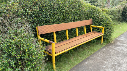 This image features a rustic park bench with a yellow metal frame and wooden slats, placed in front...